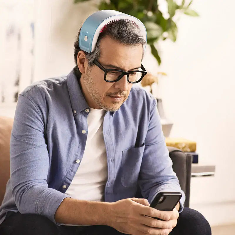 Man using LaserBand 82 hair growth device while looking at his smartphone in a modern living room.