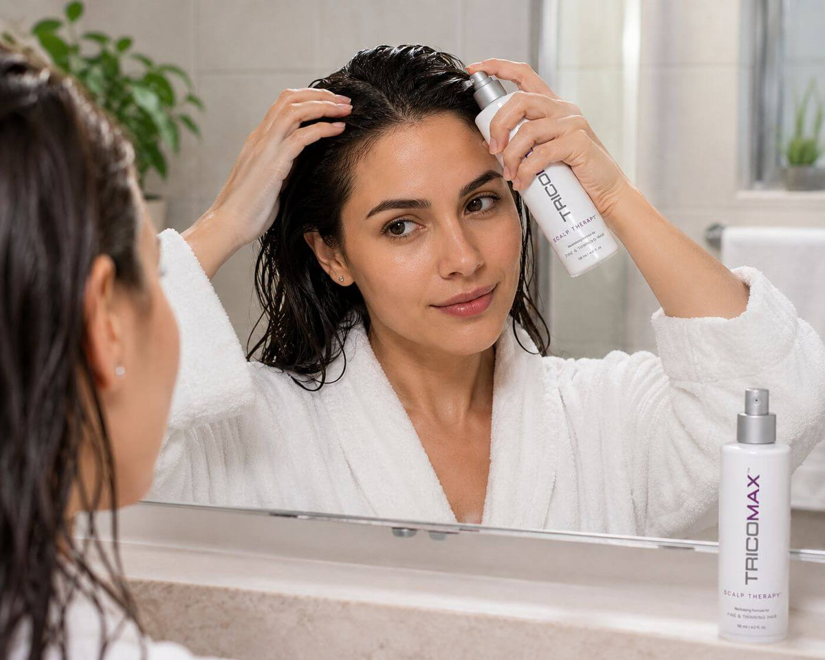 Woman applying TricoMax hair product in front of a mirror, wearing a white robe, promoting healthy hair care.